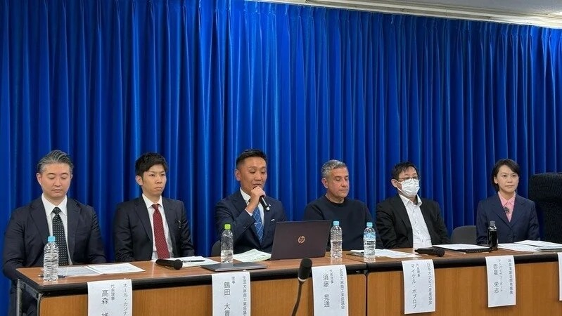 JCF記者会見の様子 - 青いカーテンを背景に業界代表者が並ぶ